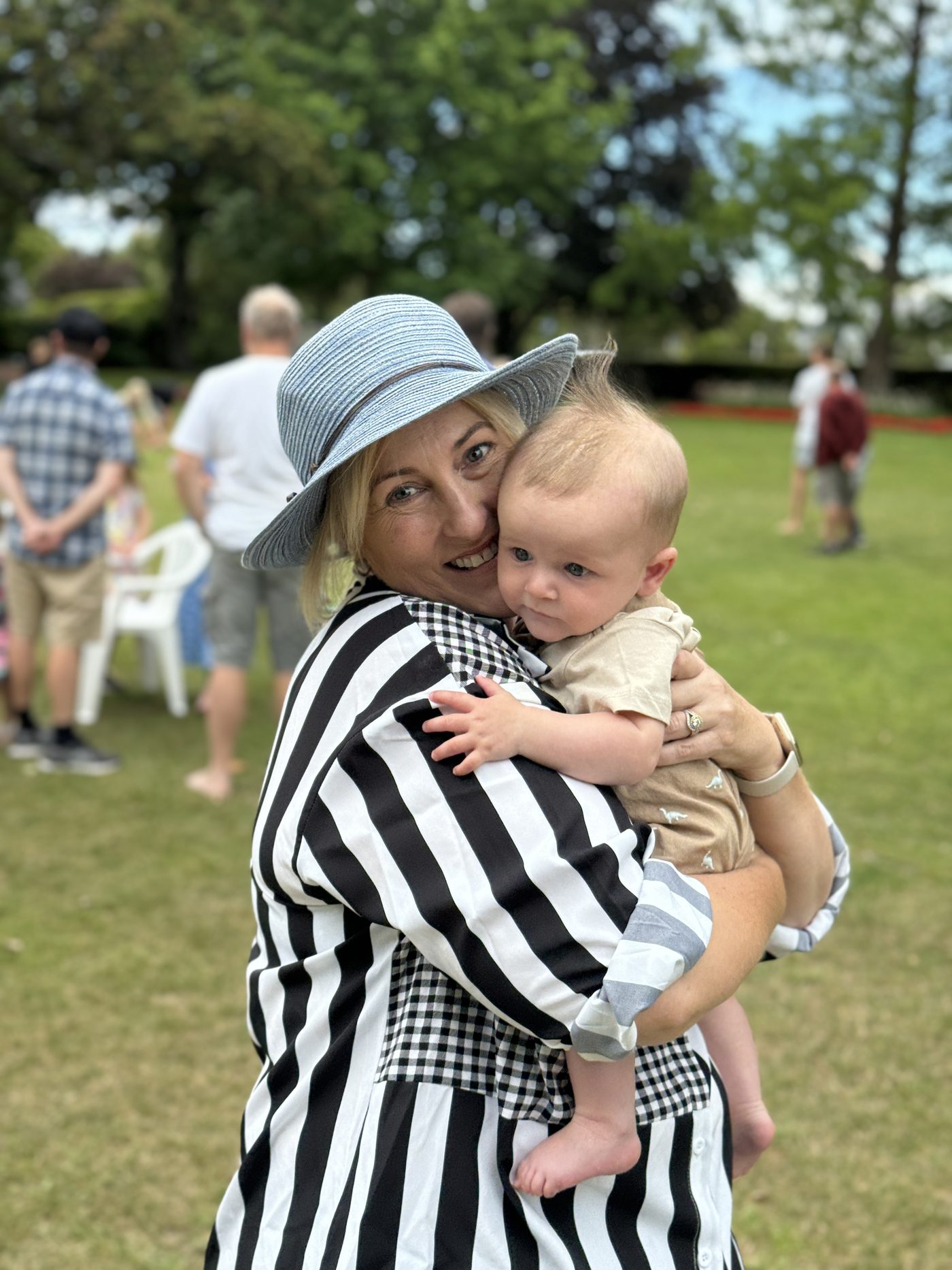 Jen in a blue sun hat at a summer picnic, cuddling her baby grandson